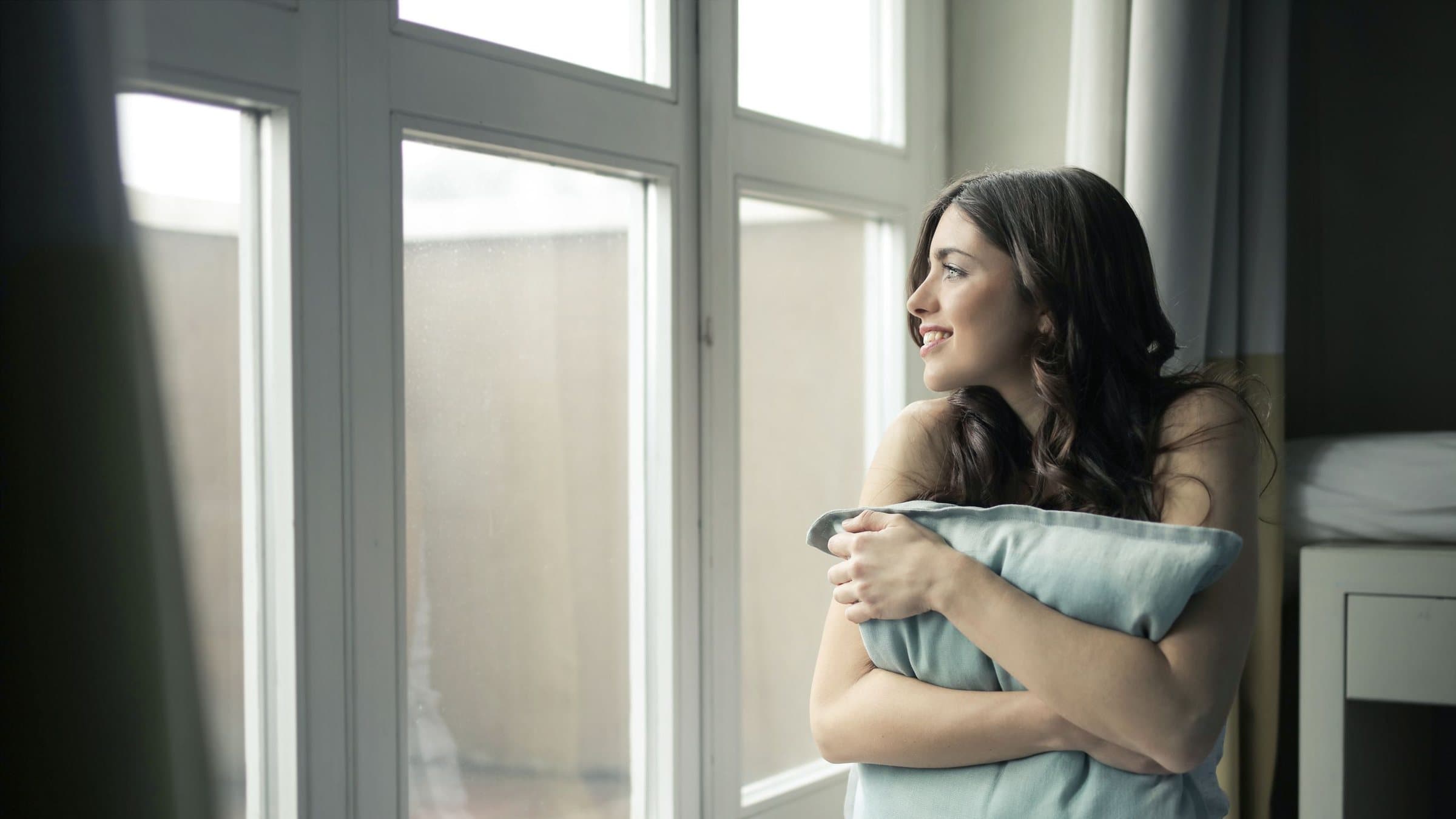 A woman sitting by a bright window holding a cushion, looking out and smiling
